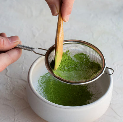 Person sifting green tea powder into a white bowl using a wooden spoon.