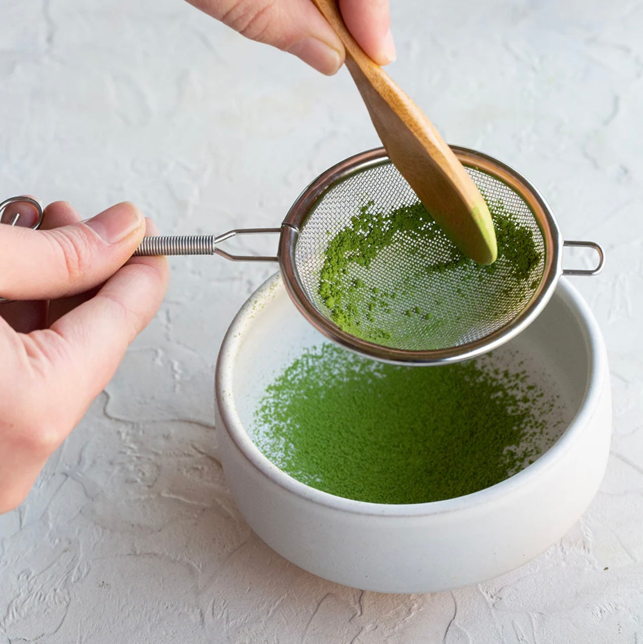 Person sifting green matcha powder into a white bowl with a wooden spoon.