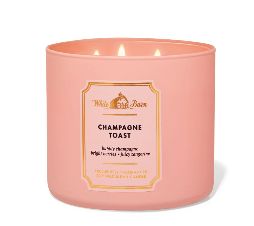 A pink 3-wick candle labeled 'Champagne Toast' from White Barn, with an image of a house and the scent marked as bright and bubbly.