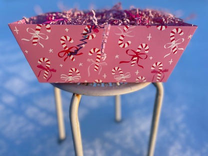 Front view of pink gift box with candy cane and bow design on a white stool in a snowy background.