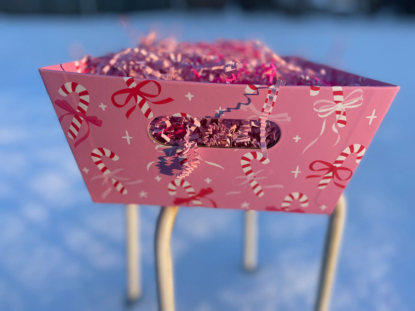 Pink decorative box with candy cane and bow pattern on a white stool in snowy background