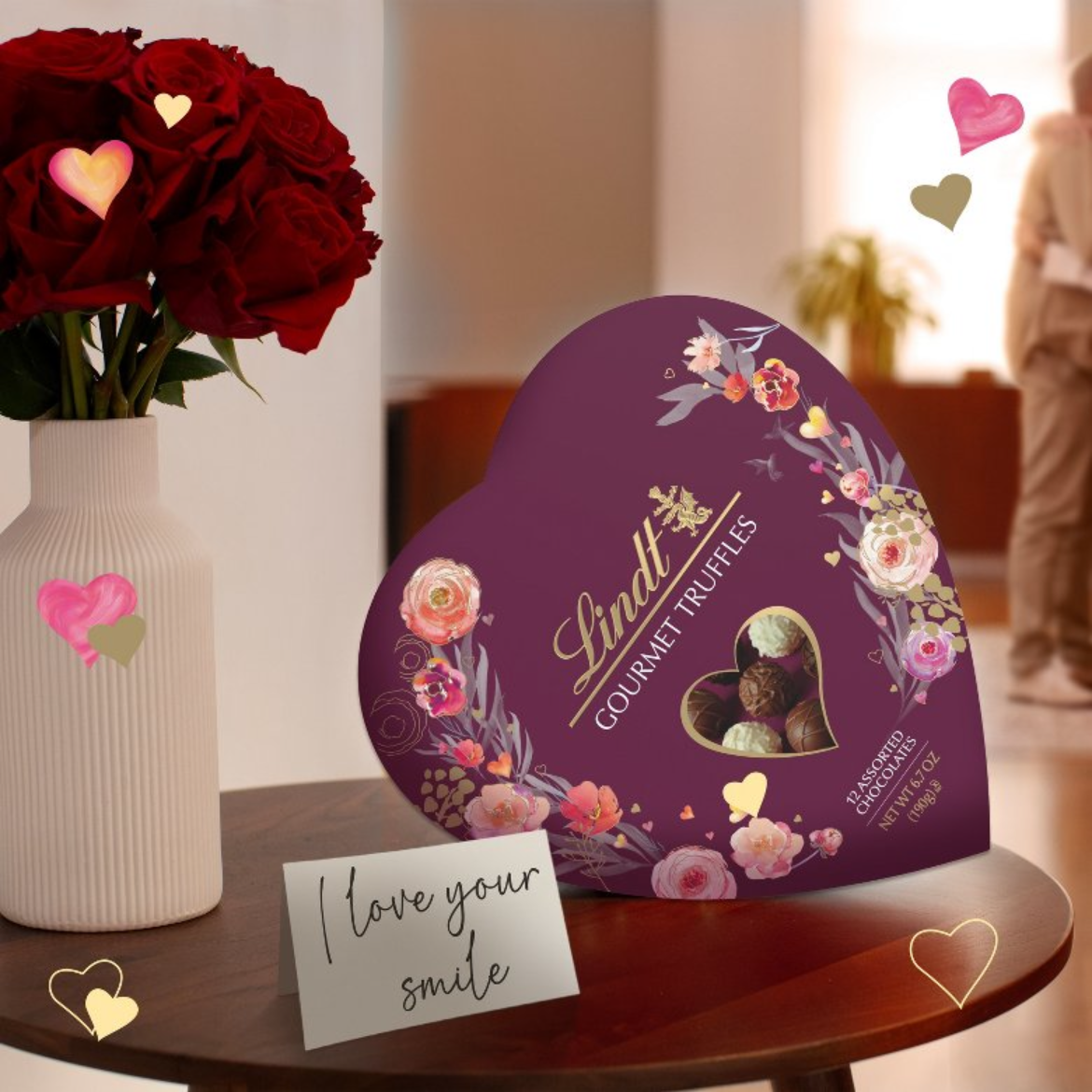 Heart-shaped Lindt chocolate box with a card on a table, surrounded by flowers.