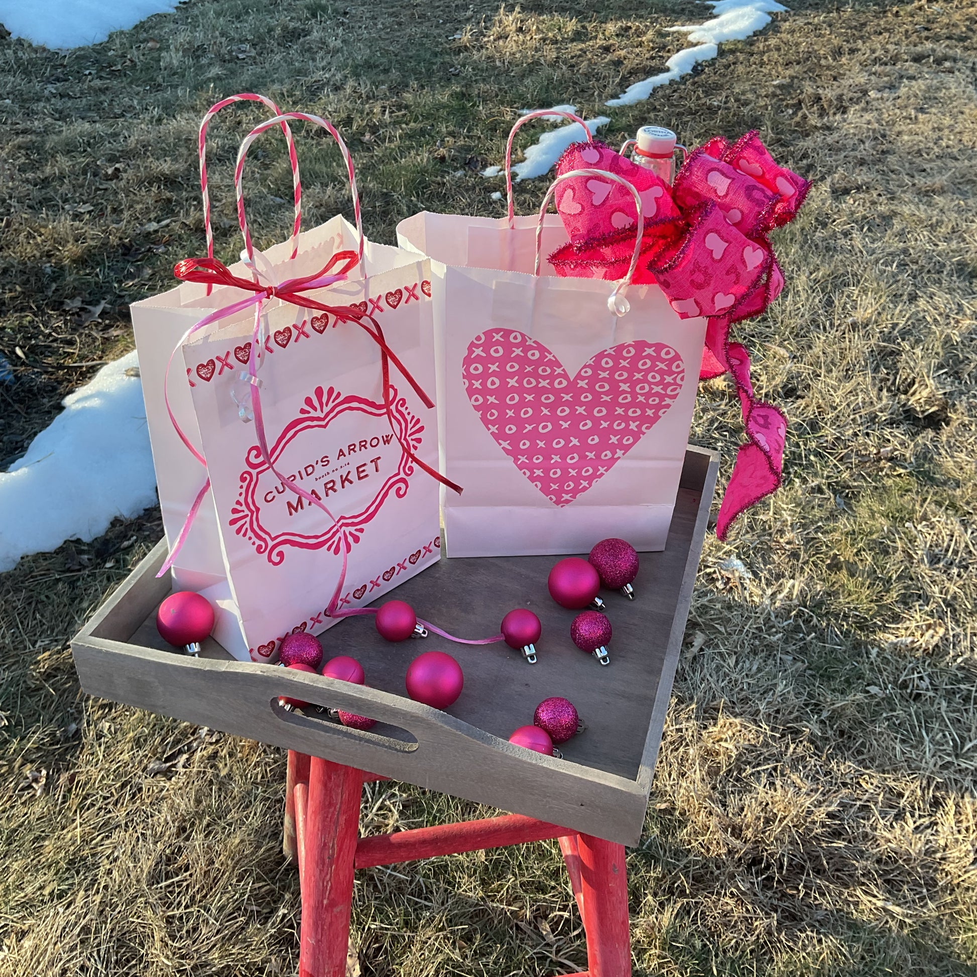 Pink gift bags with red ribbons on a wooden tray outdoors.