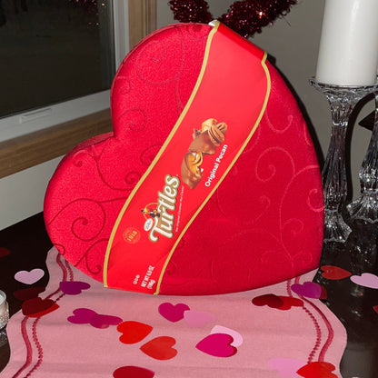 Red heart-shaped Turtles candy box on a pink tablecloth with heart patterns, candles, and decorative tinsel.
