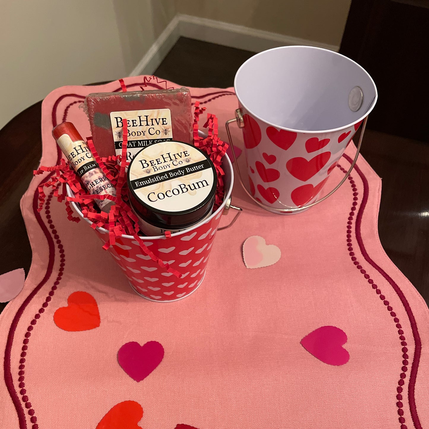 Bee Hive Body Co products in a heart-patterned bucket on a pink surface with heart designs.